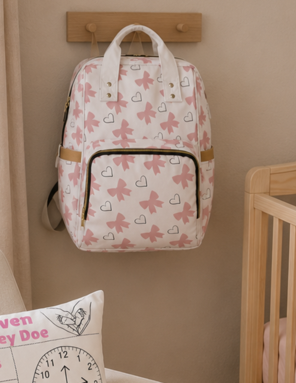 Pink and white bow and heart diaper backpack hanging on a hook in a nursery above a crib, showing the bag as part of a cozy baby room setup\.