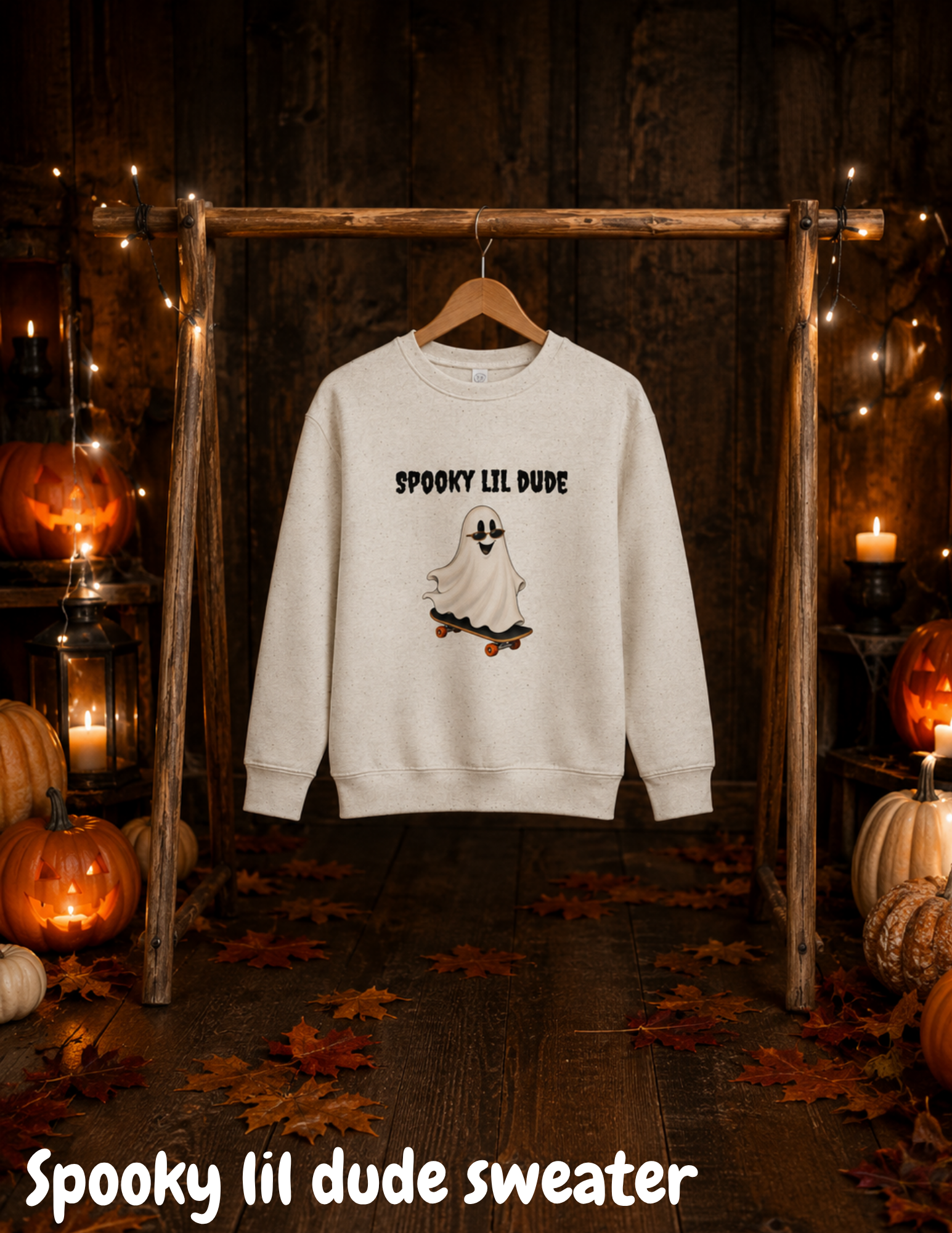 Beige Spooky Lil Dude Halloween sweater featuring a cute ghost riding a skateboard, hanging on a wooden rack surrounded by pumpkins, autumn leaves, and warm Halloween lights.